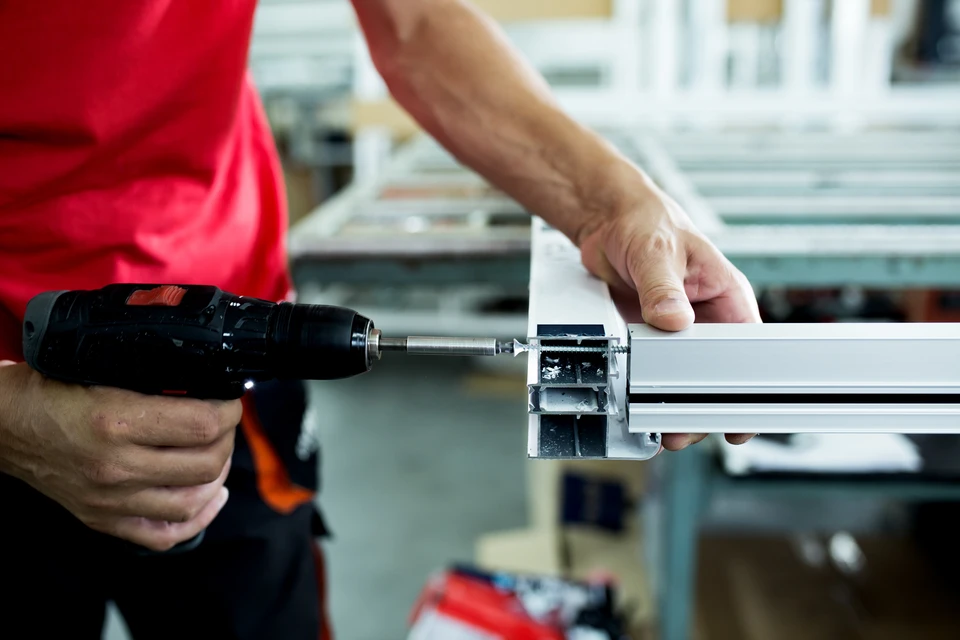 Atelier industriel avec technicien en polo rouge assemblant des profilés aluminium à l'aide d'une visseuse électrique